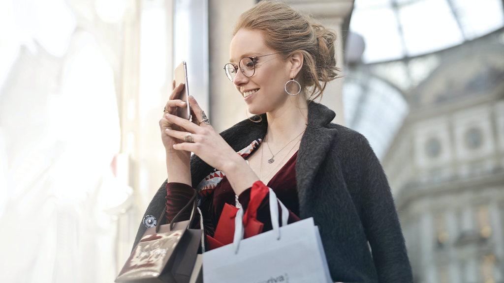 A woman checking her phone after a session of doom spending, with several shopping bags hanging off of her arms while she smiles.