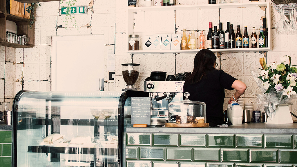 A person behind the serving counter of a restaurant decorated in white and green tiles working overtime and messing with some bottles.
