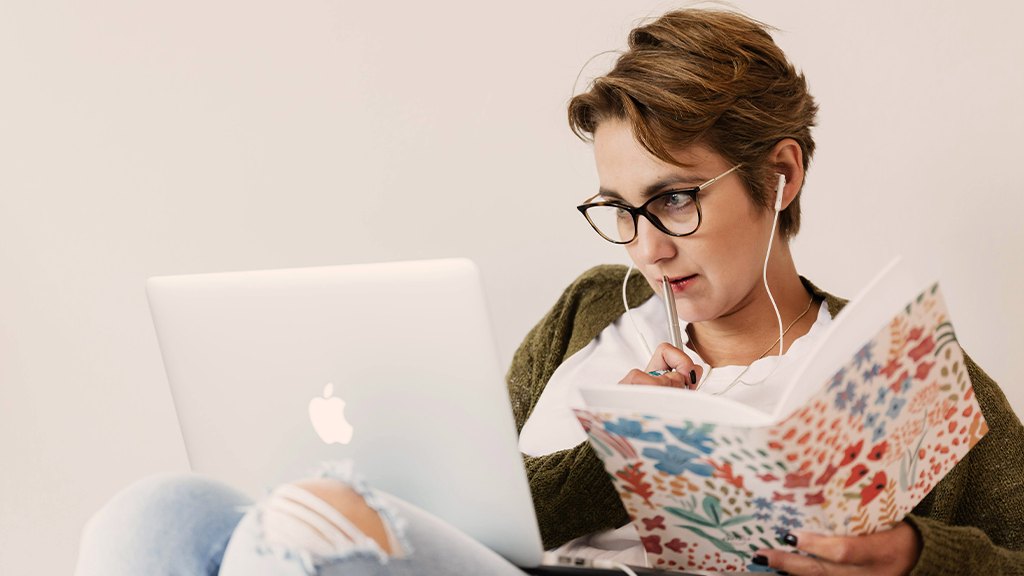A woman with short hair sits in a chair working on a laptop with a notebook open in her other hand as she reviews her social media posts.